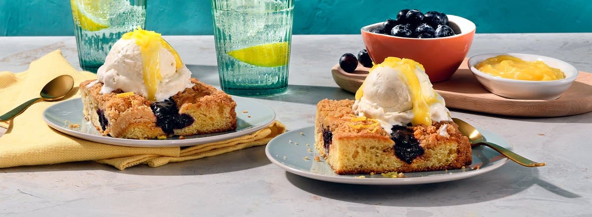Two plates of blueberry crumb cake topped with vanilla ice cream and a dollop of lemon curd. Bowl of fresh blueberries and lemon curd in the background