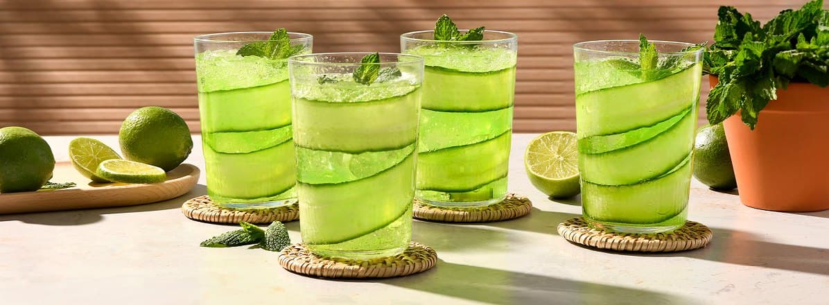Four lime rickey sodas with a cucumber ribbon lining the glass and a sprig of mint on the top. Side of sliced limes on a wooden board.