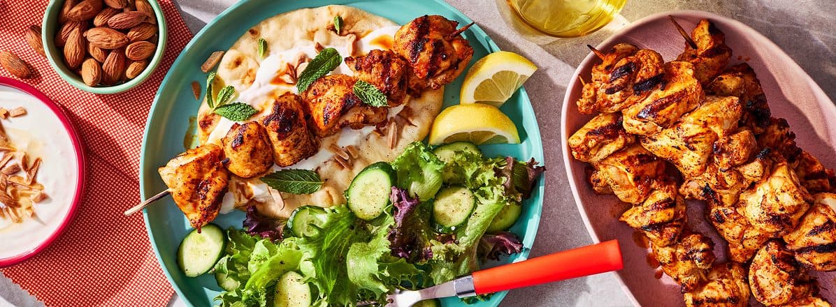 pink dinner plate with harissa grilled chicken skewer over yogurt sauce and naan bread with side green salad, and side platter of remaining chicken and harissa skewers